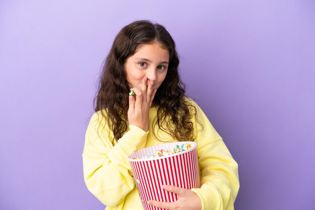 Piccola ragazza caucasica isolata su sfondo viola con in mano un grande secchio di popcorn