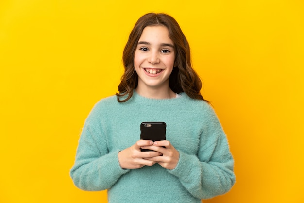 Piccola ragazza caucasica isolata sul muro giallo guardando la parte anteriore e sorridente mentre si utilizza il cellulare