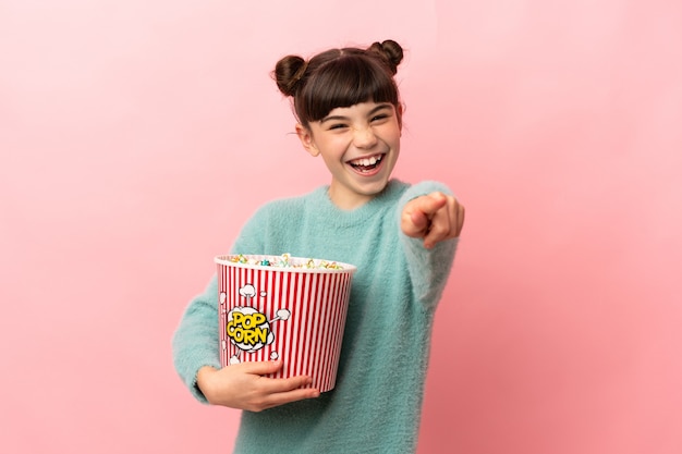 Piccola ragazza caucasica isolata tenendo un grande secchio di popcorn mentre indica la parte anteriore
