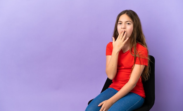 Piccola ragazza caucasica seduta su una sedia isolata su sfondo viola che sbadiglia e copre la bocca spalancata con la mano