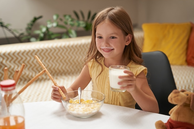 Piccola ragazza caucasica sorridente che beve latte e mangia cereali mentre fa colazione a casa