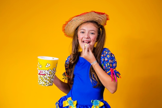 Piccola ragazza caucasica sveglia nel tipico vestito da festa junina che mangia popcorn