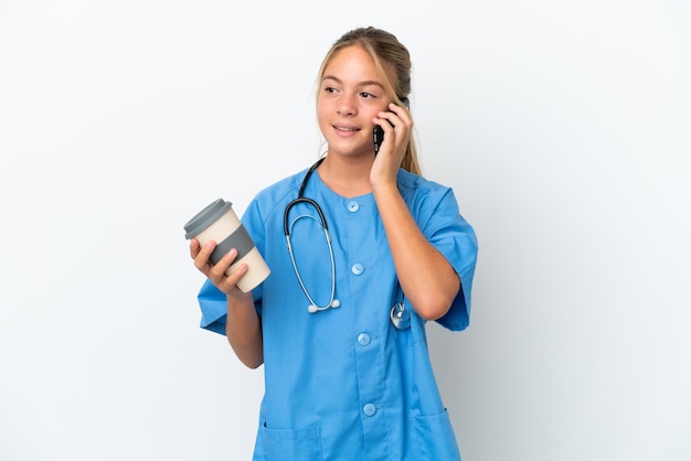 Piccola ragazza caucasica travestita da chirurgo isolata su sfondo bianco che tiene il caffè da portare via e un cellulare