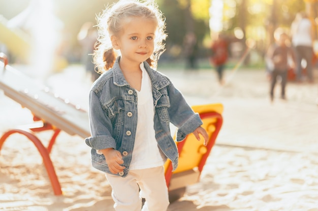 Piccola ragazza caucasica vestita con una giacca di jeans è giocata nel parco giochi in una luminosa giornata di sole