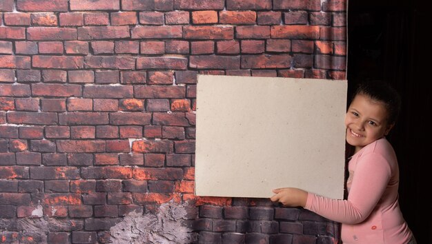 Piccola ragazza che fa pose con lettere di carta riciclata vuote davanti a un vecchio sfondo di muro di mattoni, sfondo scuro, messa a fuoco selettiva.