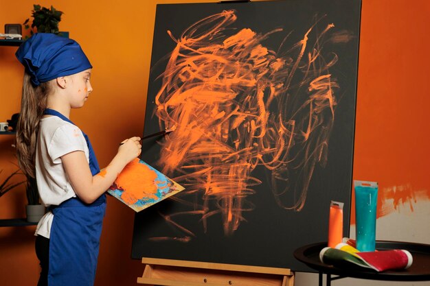 Piccola ragazza che usa il colore della vernice arancione su tela, creando un capolavoro con vassoio colormix e tavolozza aquarelle. Disegno di ispirazione alla pittura con pennello, acquerello e abilità.