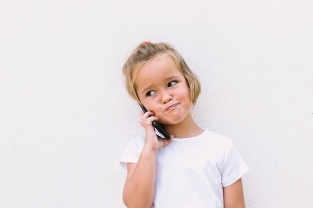 Piccola ragazza dai capelli biondi che indossa una maglietta bianca, parla al telefono cellulare, con una faccia sorridente