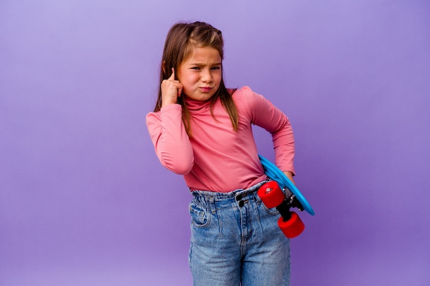 Piccola ragazza del pattinatore isolata sulle orecchie del rivestimento murale blu con le mani