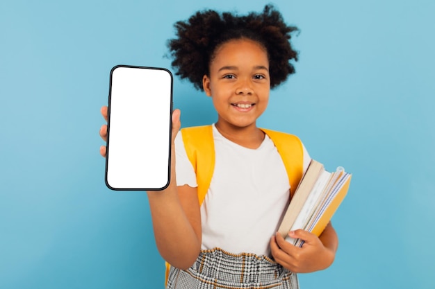 Piccola ragazza della scuola afroamericana eccitata che tiene in mano una grande cella che presenta lo schermo vuoto del telefono su sfondo blu dello studio Mockup del display dello smartphone