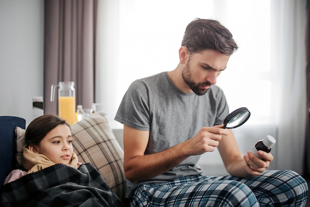 Piccola ragazza malata che si trova sul letto ed esamina la bottiglia di sciroppo la sua tenuta del papà. Lo guarda attraverso il ciclo. Il giovane sembra serio.