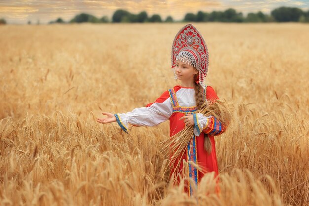 Piccola ragazza nel sarafan nazionale russo e un kokoshnik in piedi in un campo di grano dorato in estate