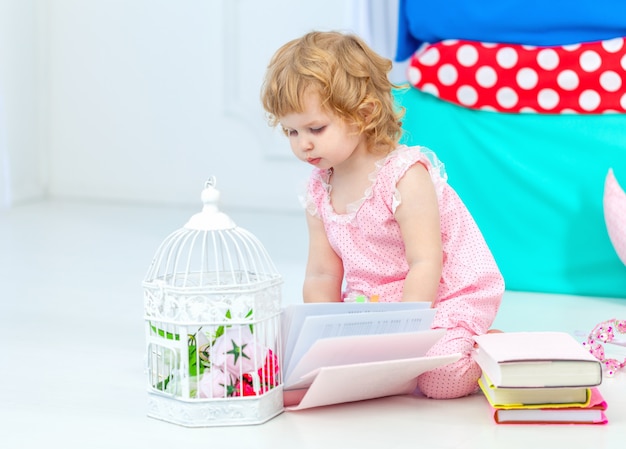 Piccola ragazza riccia sveglia in pigiami rosa che guarda il libro che si siede sul pavimento nella camera da letto dei bambini.