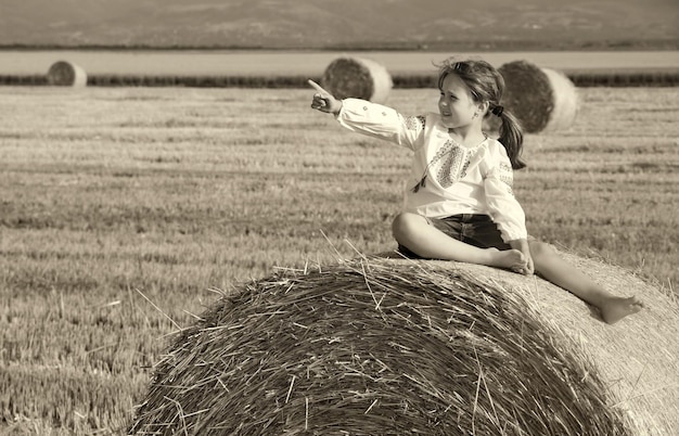 Piccola ragazza rurale sulla paglia dopo il campo di raccolta con balle di paglia