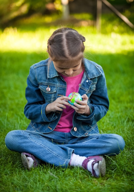 Piccola ragazza seduta sull'erba al parco ed esplorando il globo