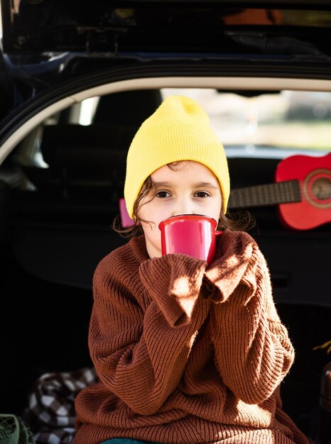 Piccola ragazza sorridente carina con berretto giallo seduta nel bagagliaio di un'auto aperta e bevendo cacao Bambino che riposa con la sua famiglia nella natura vicino all'acqua Stagione autunnale