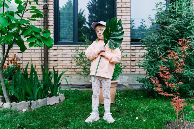 Piccola ragazza sorridente che tiene una foglia di monstera davanti alla casa nel giardino verde