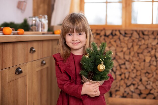 Piccola ragazza sorridente in abito rosso che mostra un piccolo abete di Natale decorato in una pentola in casa