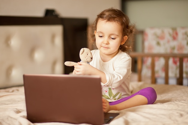 Piccola ragazza sveglia che si siede sul letto e che per mezzo di un taccuino del computer portatile della compressa digitale. Chiamate online amici o genitori.