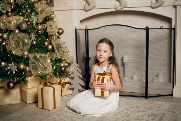 Piccola ragazza sveglia in vestito che si siede vicino all'albero di Natale e che tiene i regali.