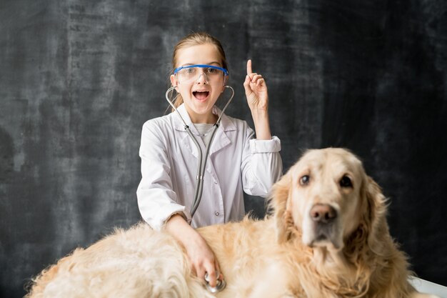 Piccola ragazza veterinaria sveglia in occhiali protettivi che esamina il cane di labrador con lo stetoscopio e che indica verso l'alto