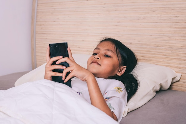 Piccola ragazzina carina che guarda un video sullo smartphone con la faccia sorridente da sola sul letto