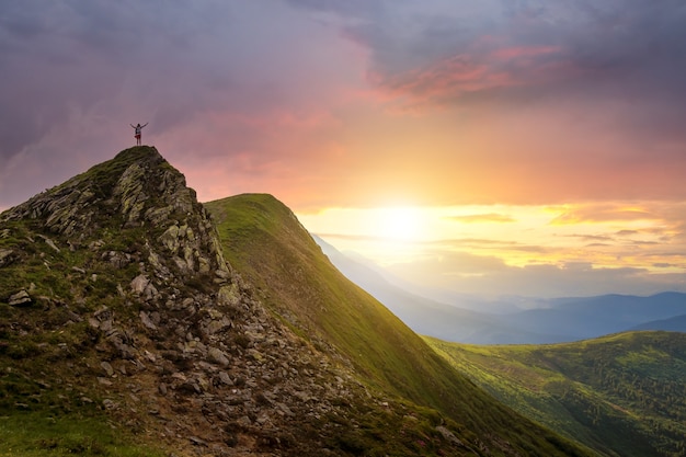 Piccola silhouette di un escursionista in piedi con le braccia alzate sulla cima di una montagna rocciosa al tramonto.