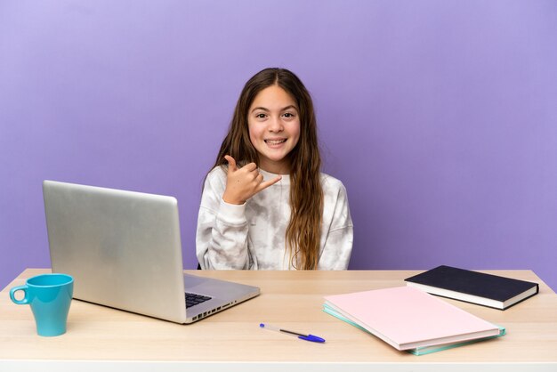 Piccola studentessa in un posto di lavoro con un computer portatile isolato su sfondo viola che fa il gesto del telefono. Richiamami segno