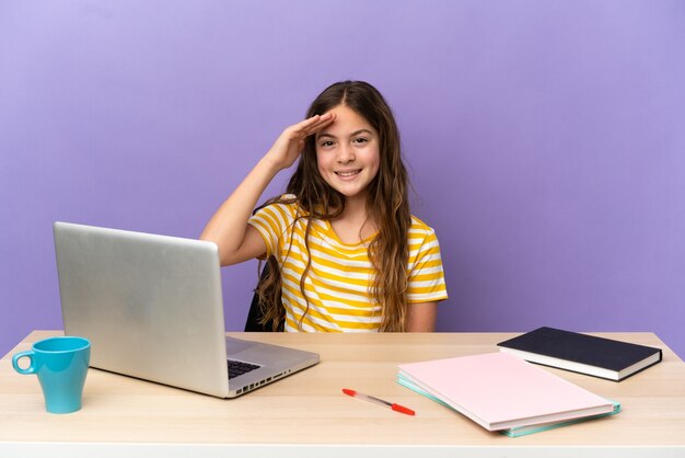 Piccola studentessa in un posto di lavoro con un computer portatile isolato su sfondo viola che saluta con la mano con espressione felice