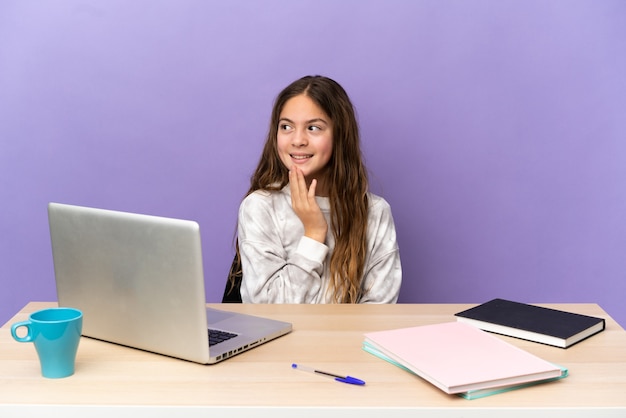Piccola studentessa in un posto di lavoro con un laptop isolato su sfondo viola che guarda in alto sorridendo