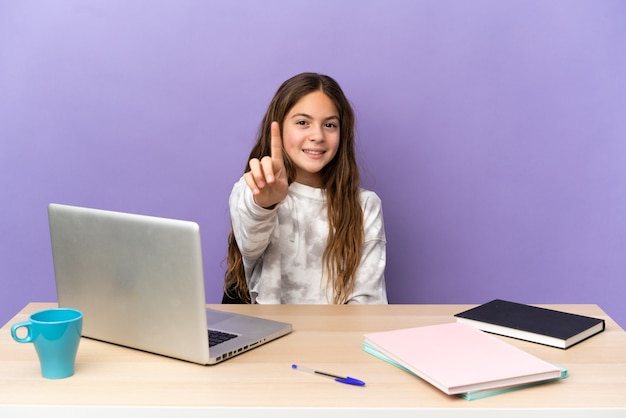 Piccola studentessa in un posto di lavoro con un laptop isolato su sfondo viola che mostra e solleva un dito