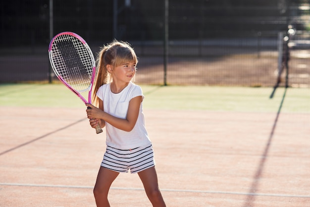 Piccola tennista in uniforme bianca concentrandosi e concentrandosi sull'allenamento prima della partita. Sfondo di luce solare.