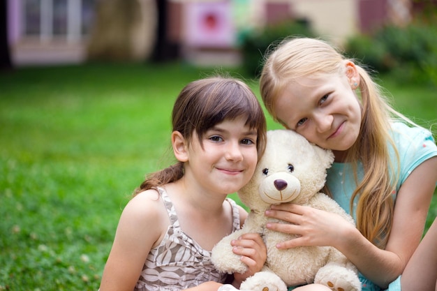 Piccole ragazze graziose amiche sedute sul prato con il suo orsacchiotto giocattolo. Felice estate