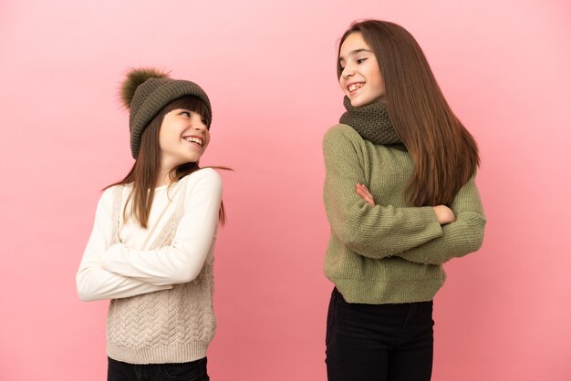 Piccole sorelle che indossano abiti invernali isolati su sfondo rosa guardando sopra la spalla con un sorriso