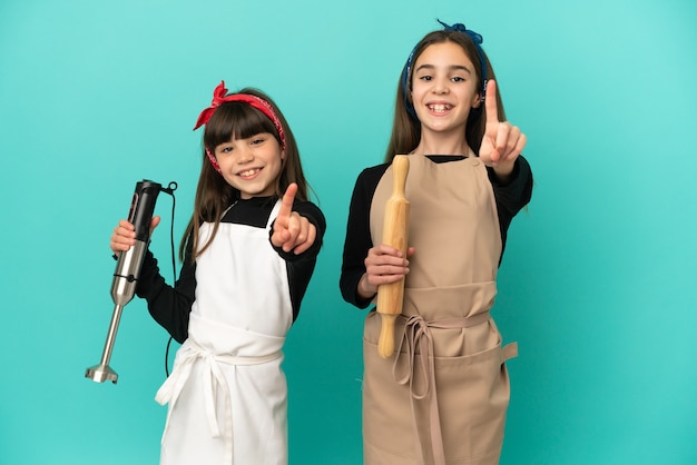 Piccole sorelle cucinare a casa isolato su sfondo blu che mostra e alzando un dito