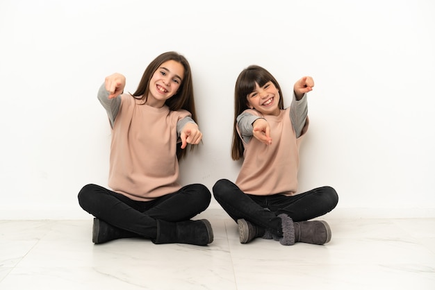 Piccole sorelle seduti sul pavimento isolato su sfondo bianco punta il dito contro di te mentre sorridono