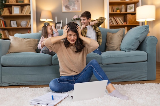 Piccoli bambini cattivi che fanno rumore e distraggono la madre freelance che cerca di concentrarsi sul portatile e stringe la testa che lavora al tavolo nel salotto luminoso