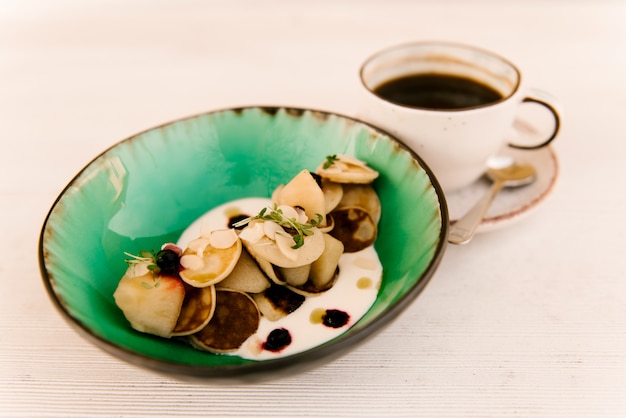 Piccoli pancake in un piatto con le bacche e la panna acida casalinga con una tazza di caffè su un fondo bianco, fuoco selettivo