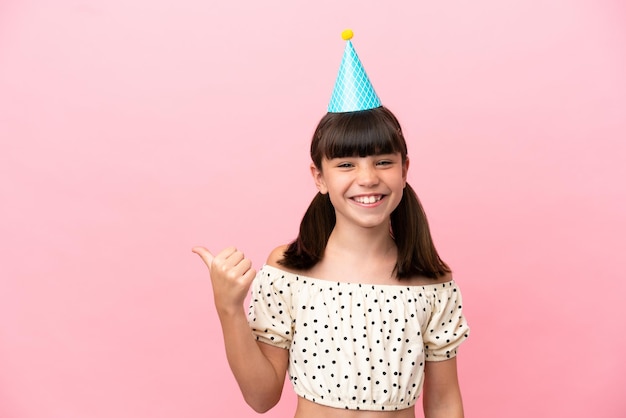 Piccolo bambino caucasico con cappello di compleanno isolato su sfondo rosa che punta al lato per presentare un prodotto