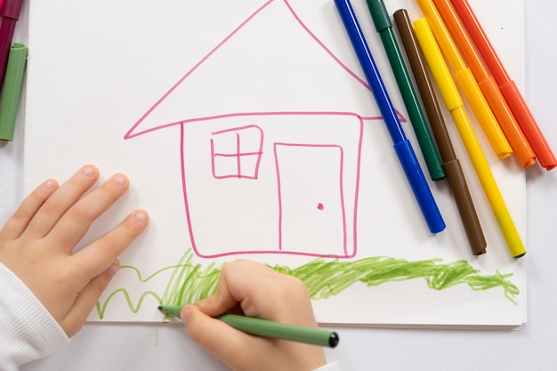 Piccolo bambino che disegna con penne felttip una casa con l'erba in un quaderno di disegni