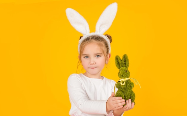 piccolo bambino di pasqua in orecchie da coniglio che tiene giocattolo su sfondo giallo.