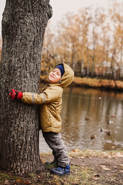 Piccolo bambino felice di quattro anni, che ha stretto gli occhi chiusi, in abiti autunnali abbraccia un tronco d'albero durante una passeggiata nel parco autunnale vicino allo stagno
