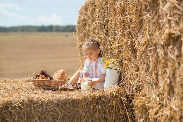piccolo bambino felice in abiti popolari si siede su un campo di grano con un barattolo di latte fresco.