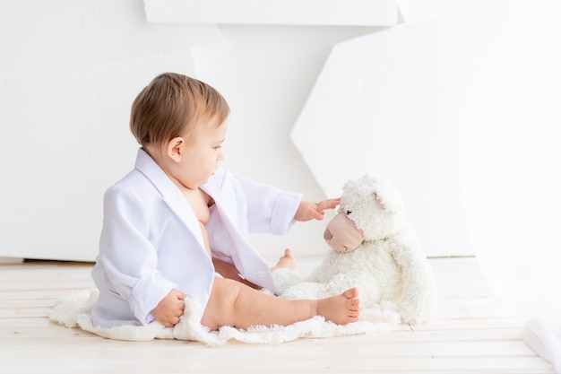 Piccolo bambino grasso in una giacca bianca e pannolini gioca con un orsacchiotto