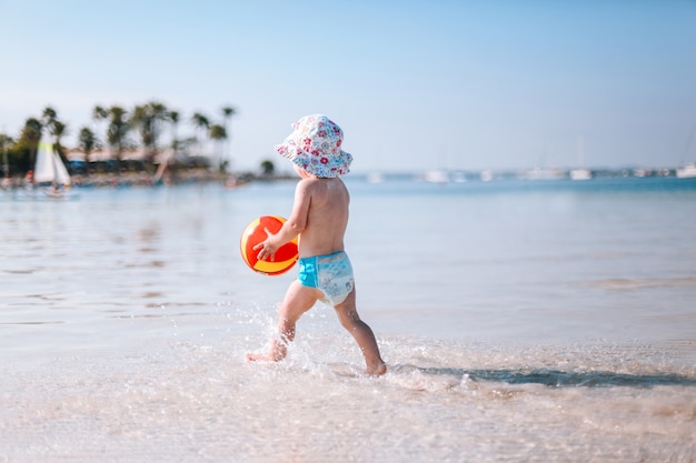 Piccolo bambino riccio sveglio gioca con la palla variopinta sulla spiaggia. Bambina che cammina sull'acqua in riva al mare.