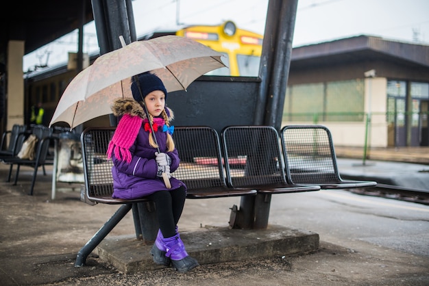 Piccolo bambino seduto sulla piattaforma con un ombrello su una stazione ferroviaria