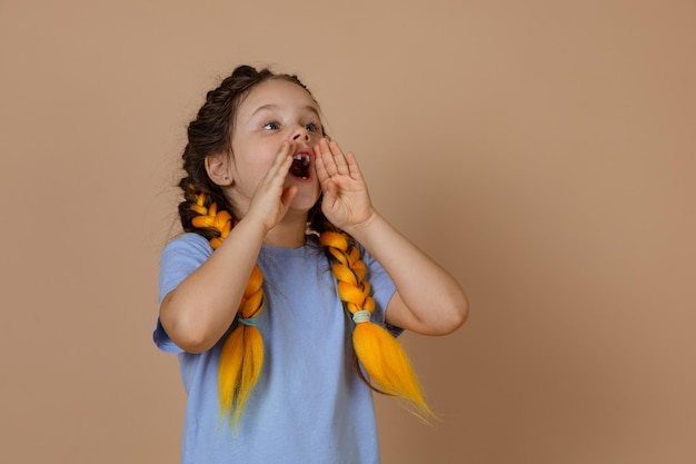 Piccolo bambino spaventato che urla tenendosi per mano con la bocca aperta chiedendo aiuto con trecce kanekalon di colore giallo che guarda in alto su sfondo beige indossando una maglietta blu