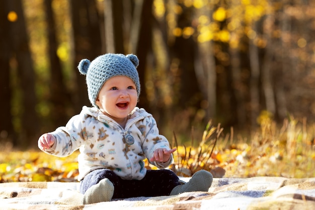 Piccolo bambino sveglio in buffo cappello con orecchie e felpa con cappuccio seduto nel parco in una giornata di sole autunnale