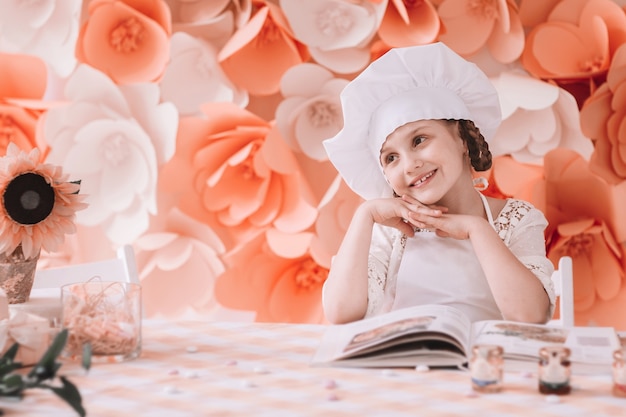 Piccolo capo in cucina. Affascinante bambina in un cappello da capo sta a un tavolo da pranzo pronto a cucinare una cena