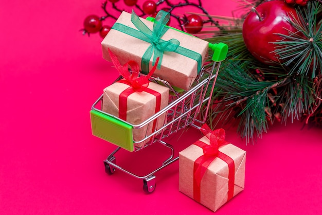 Piccolo carrello della spesa con scatole regalo sulla superficie rosso-rosa con albero di Natale