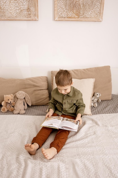 Piccolo e simpatico ragazzo caucasico che legge un libro sul letto di casa. Interni e vestiti nei colori della terra naturale. Ambiente accogliente. Il bambino legge una fiaba.